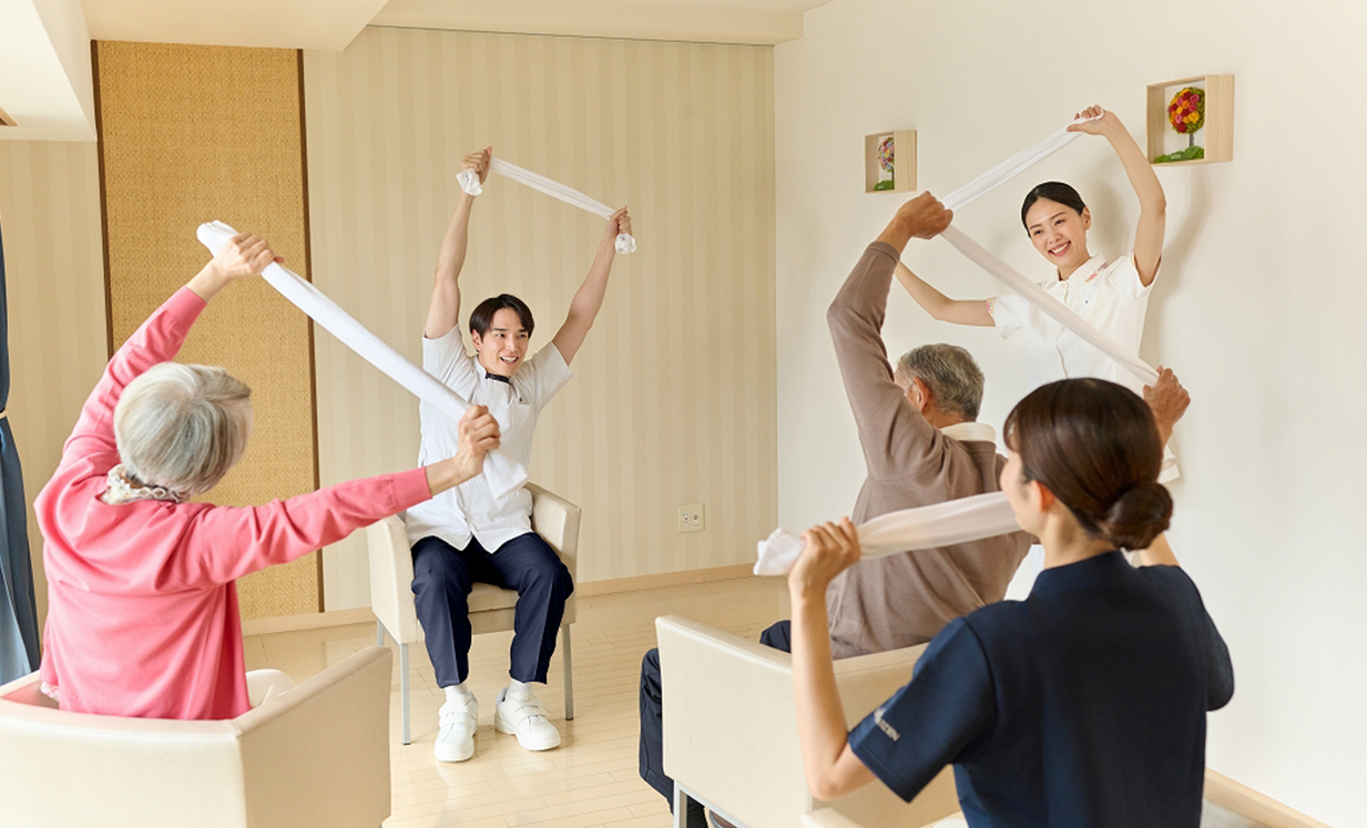 介護職員と入居者による機能訓練。白いタオルを使った集団レクリエーション活動